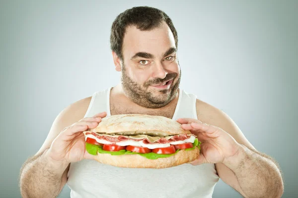 Толстый смешной человек ест огромный бутерброд, изолированный на серый — стоковое фото