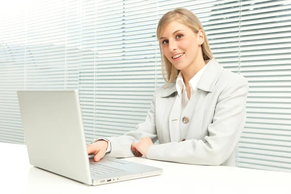 Donna d'affari bionda sorridente con laptop vicino alla finestra veneziana cieca — Foto Stock