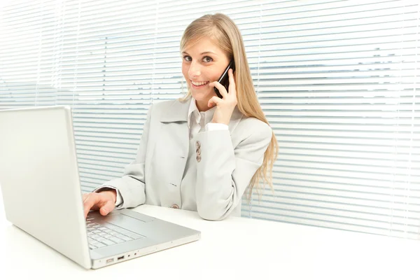 Donna d'affari bionda sorridente con computer portatile parlare al telefono vicino alla finestra veneziana cieca — Foto Stock