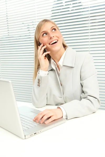 Donna d'affari bionda sorridente con computer portatile parlare al telefono vicino alla finestra veneziana cieca — Foto Stock
