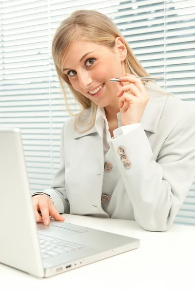 Mulher de negócios loira sorridente com laptop perto de janela cega veneziana — Fotografia de Stock