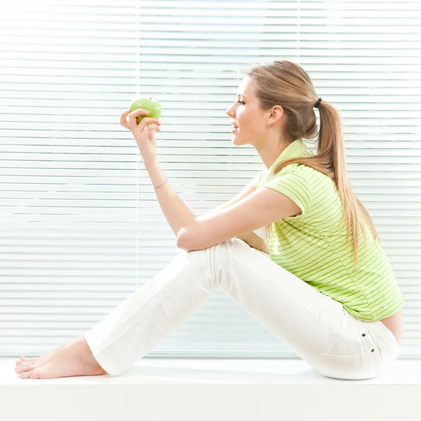 젊은 금발 미인 블라인드 창 배경 녹색 사과 먹으십시오 — 스톡 사진