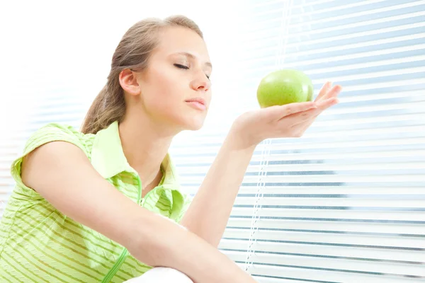Young beautiful blond woman eat green apple with venetian blind window background — Stock Photo, Image