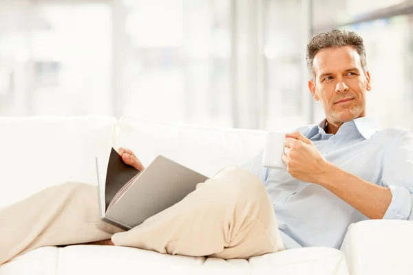 Bell'uomo d'affari leggere e bere una tazza di tè sul divano di casa — Foto Stock