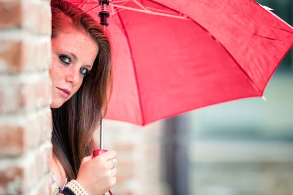 Fiatal vöröshajú lány piros esernyő, egy utca-és városrészlet — Stock Fotó