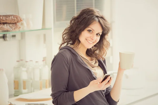 Joven mujer sonriente con taza de té y teléfono en la cocina — Foto de Stock