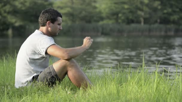 Jongeman zit op gras neem foto met smartphone op de oever van het meer — Stockvideo