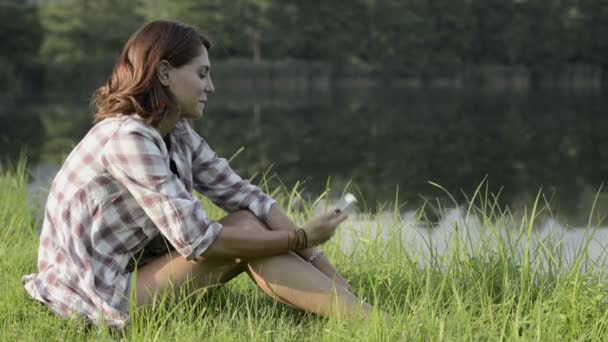 Молодая женщина, сидящая на траве, фотографируется со смартфоном на берегу озера — стоковое видео