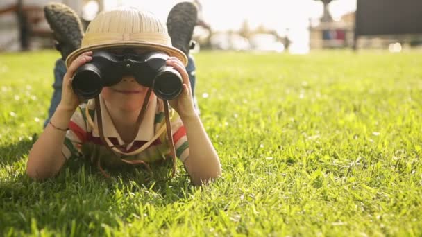 Child wearing pith helmet is playing in summer day with binoculars lying down — Stock Video