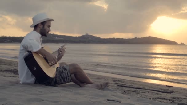 Unga vackra man sitter utomhus på stranden spela gitarr — Stockvideo