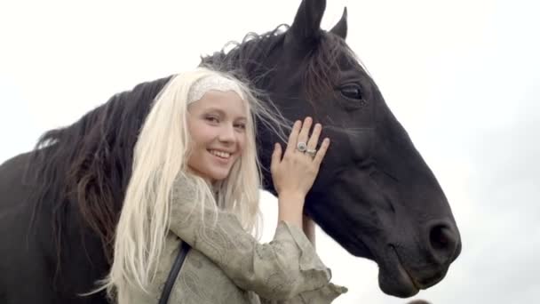 Blondynka młoda kobieta uśmiecha się w pociągnięcia i przytula czarny koń — Wideo stockowe