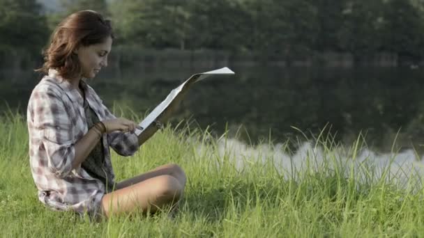 Young woman in nature seated on grass look at map on lake shore — Stock Video