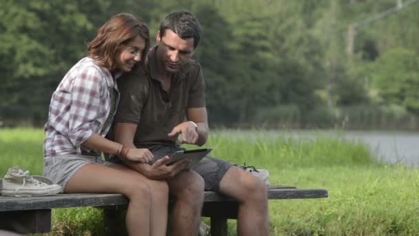 Pareja joven en la naturaleza mira la tableta sentada en el embarcadero del lago — Vídeos de Stock