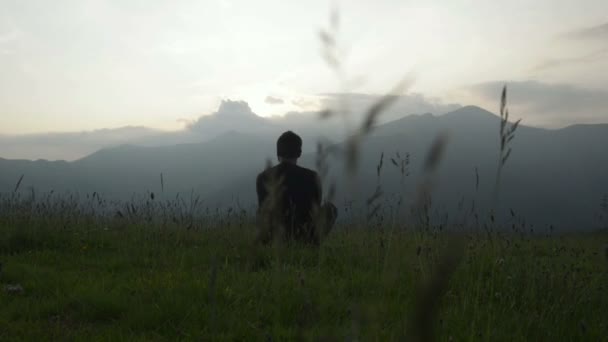 Joven sentado en la hierba está mirando al panorama al atardecer — Vídeos de Stock