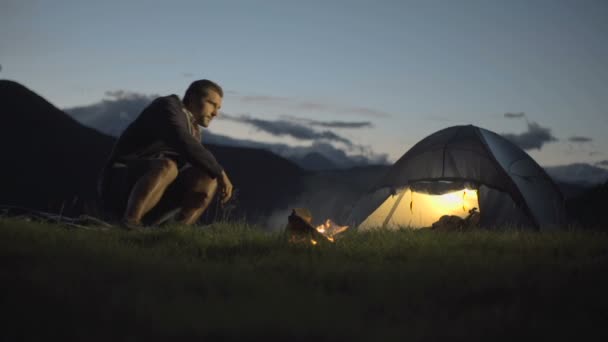 Hombre joven entra y se calienta con fuego de campamento en la naturaleza montaña al aire libre — Vídeo de stock