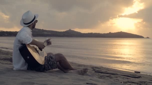 Joven hermoso hombre sentado al aire libre en la orilla toca la guitarra — Vídeos de Stock