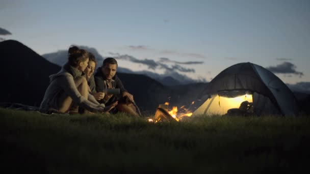 Grupo de três amigos conversando, sorrindo e aquecendo com fogo de acampamento na natureza — Vídeo de Stock