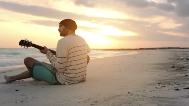 Joven hombre hermoso toca la guitarra en la playa del océano junto al mar al amanecer — Vídeos de Stock