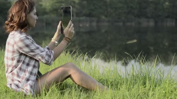 Молодая женщина, сидящая на траве, фотографируется с табличкой на берегу озера — стоковое видео