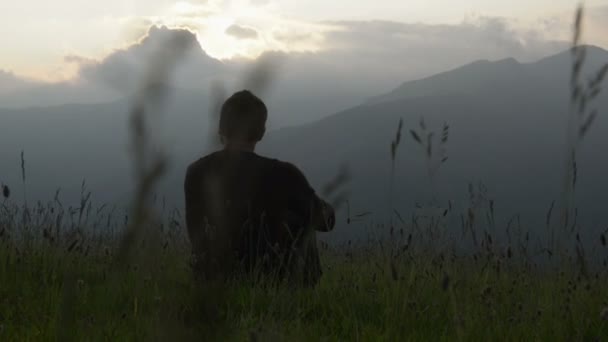 Joven sentado en la hierba está mirando al panorama al atardecer — Vídeos de Stock