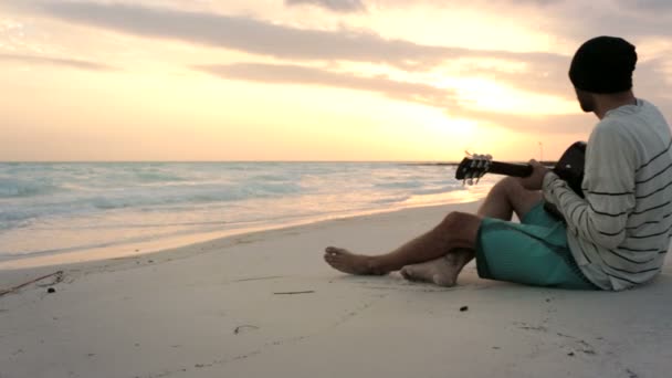 Joven hombre hermoso toca la guitarra en la playa del océano junto al mar al amanecer — Vídeos de Stock