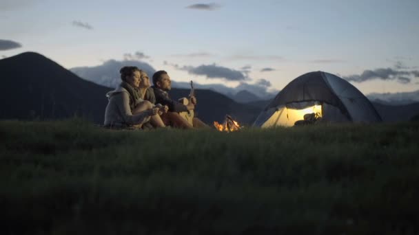 Gruppo di tre amici suonare la chitarra e cantare al fuoco campo in montagna natura — Video Stock