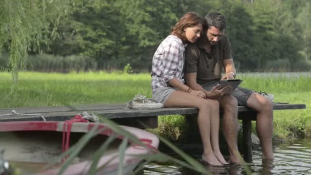Пара смотрит на планшет, сидящий на причале озера — стоковое видео
