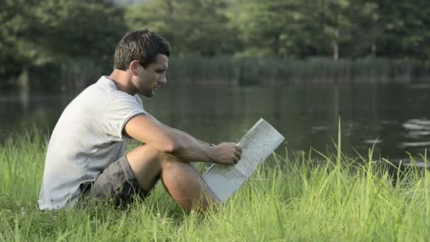 Jeune homme dans la nature assis sur l'herbe regarder la carte sur la rive du lac — Video