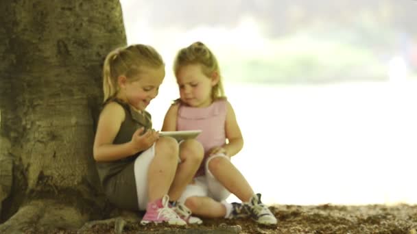 Hermanas rubias felices están leyendo cuentos de hadas en la tableta en el parque de la ciudad — Vídeos de Stock