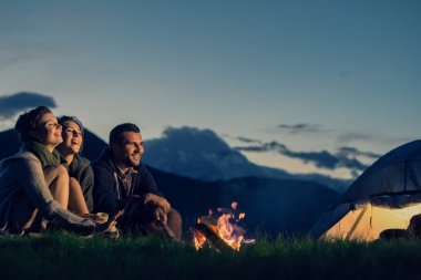 Three friends camping with fire on mountain at sunset clipart