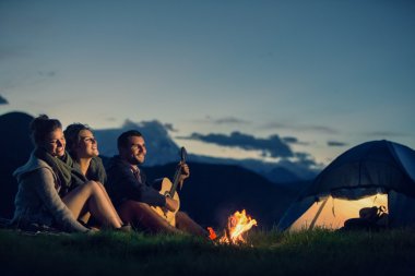 Three friends camping with fire on mountain at sunset clipart