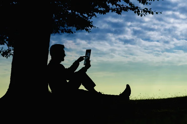 Silhueta homem sentado sob a árvore com comprimido no dia nublado ao ar livre — Fotografia de Stock