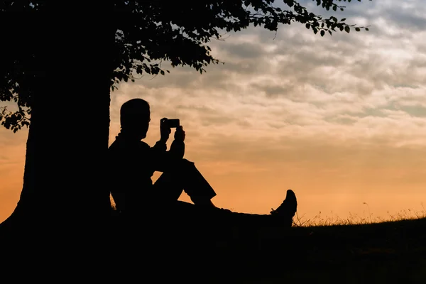 Silueta muž sedí pod stromem s telefonem na zamračený den venkovní — Stock fotografie