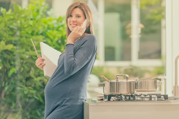 Беременная брюнетка говорит по телефону на кухне — стоковое фото