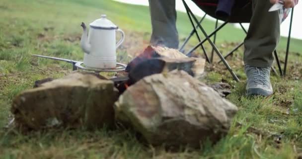 Mladý muž sedící před ohněm. Muž krájí nožem prut. Káva na ohni. — Stock video