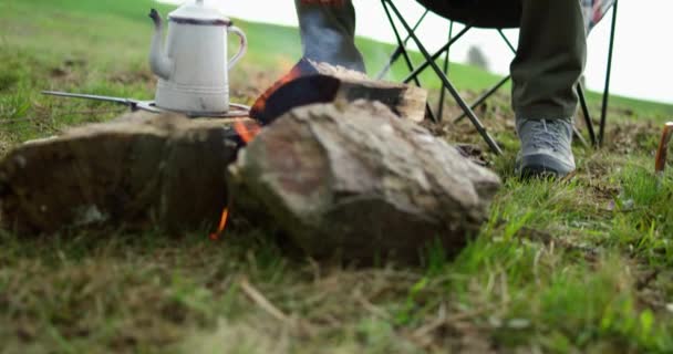 Een jongeman voor het kampvuur. Een man die met een mes een takje snijdt. Koffie op het vuur. — Stockvideo