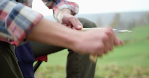 Close-up van de handen van de mens snijden met mes een takje, zittend op campingstoel. Bewolkte lucht op de achtergrond — Stockvideo