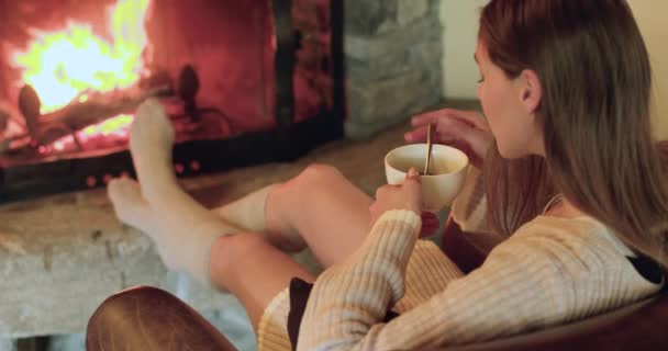 Hermosa mujer sentada calentando sus pies cerca de la chimenea con calcetines de lana, bebiendo una taza de café caliente. Acogedor material de archivo en casa, concepto de Navidad, concepto pasado de moda. — Vídeo de stock