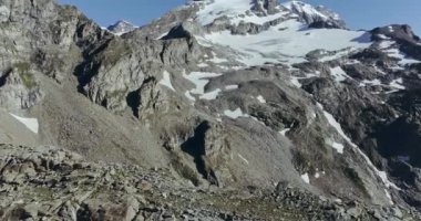 Kayalık ve karlı dağ zirvesi, yerdeki kayalar ve dağ sırasının arkasındaki mavi gökyüzü.