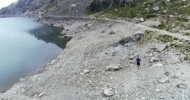 Um homem a correr numa estrada rochosa na cave de uma montanha rochosa. Lago na costa da montanha. — Vídeo de Stock