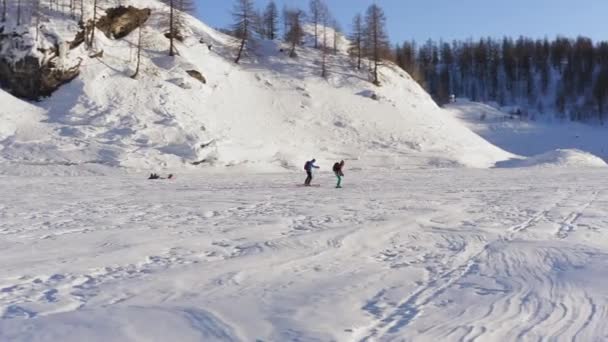 Letecký pohled na lidi procházející zasněženou oblastí na slunci. Stín hor pokrývá část oblasti. Horské pásmo v pozadí. — Stock video