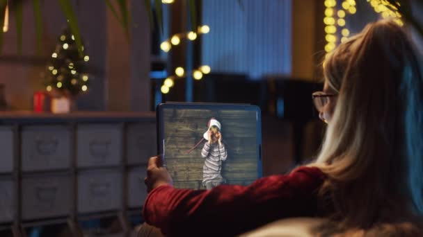 Dijital tablet tutan genç bir kadının arka plan görüntüsü. Kız kardeşiyle uzaktan çekilmiş bir videoda konuşan, gülümseyen bir kadın. Noel toplarıyla oynayan. Arka planda Noel ağacı, süslemeler ve ışıklar — Stok video