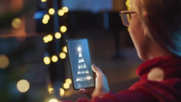 Widok młodej kobiety trzymającej telefon komórkowy. Kobieta ogląda, uśmiecha się na kartkę świąteczną od przyjaciół, rodziców.W tle dekoracje świąteczne i światło. Boże Narodzenie strój w domu. — Wideo stockowe