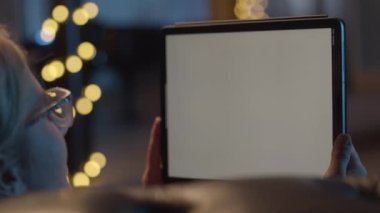 Kanepeye uzanmış dijital tabletinde film izleyen bir kadın. Arka planda Noel ağacı, süslemeler, ışık. Noel tatili boyunca evde dinlenmek.