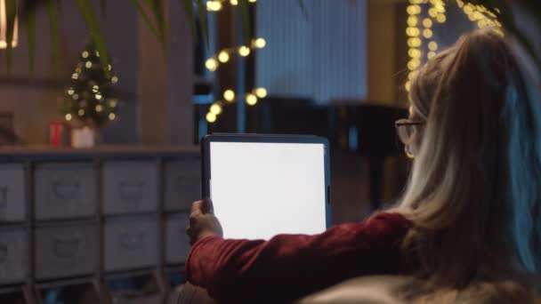 Vista posterior de una mujer joven sosteniendo una tableta digital.Mujer hablando, sonriendo, riendo en un videollamada con sus amigos, padres. En el fondo Árbol de Navidad, decoraciones y luz. — Vídeos de Stock
