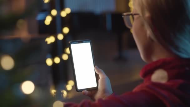 Baksidan av ung kvinna som håller mobiltelefon. Kvinnan pratar, ler, skrattar i en video ringer med sina vänner, föräldrar. I bakgrunden julpynt och ljus. Julkläder hemma. — Stockvideo