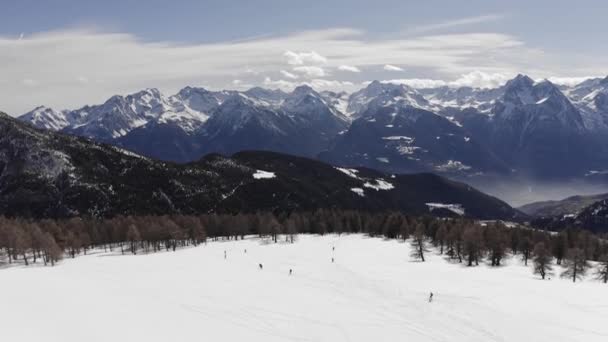 Vue aérienne du sommet du groupe de skieurs descendant la montagne, fuyant la forêt de pins, descendant la pente ensoleillée. Chaîne de montagnes enneigée en arrière-plan. au ralenti — Video