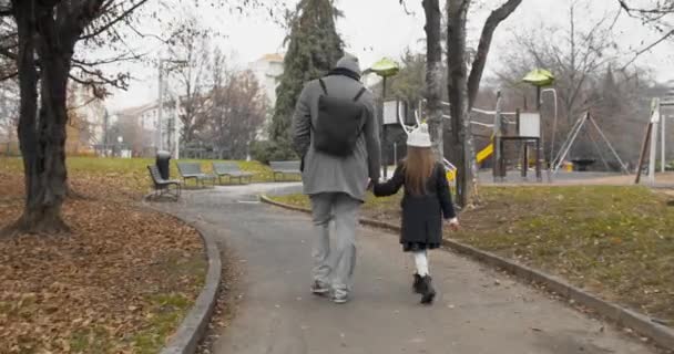 Widok ojca i jego dziewczynki idących do parku. Kobieta dziecko z białą maską i ojciec ręka w rękę chodzenie do parków przejażdżki w pochmurną jesienną podróż — Wideo stockowe