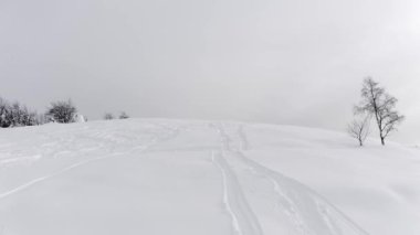 Kışın ve sisli bir yolculukta kar üzerinde kayak izleri olan karlı dağ zirvesinin havadan görünüşü. Yan taraftaki ağaçlar