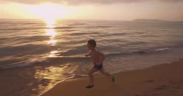 Kind loopt op het strand bij zonsondergang. Jongen heeft plezier op zand en zee. op de achtergrond gouden zon schijnt en zomer dorp met boom — Stockvideo